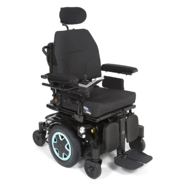 The image shows a power wheelchair with a black seat, armrests, and headrest. The chair has a robust frame with teal accents on the wheels. The wheelchair features footrests and various controls on the side, designed for individuals with mobility impairments.